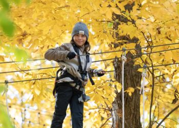 A young boy with long brown hair, wearing a gray knit "COAL" beanie and a neutral-toned fleece jacket over a black zip-up, smiles at the camera while navigating an aerial ropes course. He is wearing fingerless black gloves and is securely attached to a safety harness and belay system. He stands on a thick cable, holding onto a parallel handrail cable with both hands. The background is a vibrant, out-of-focus canopy of bright yellow autumn leaves and the thick trunk of a deciduous tree, illuminated by soft daylight.