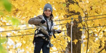 A young boy with long brown hair, wearing a gray knit "COAL" beanie and a neutral-toned fleece jacket over a black zip-up, smiles at the camera while navigating an aerial ropes course. He is wearing fingerless black gloves and is securely attached to a safety harness and belay system. He stands on a thick cable, holding onto a parallel handrail cable with both hands. The background is a vibrant, out-of-focus canopy of bright yellow autumn leaves and the thick trunk of a deciduous tree, illuminated by soft daylight.