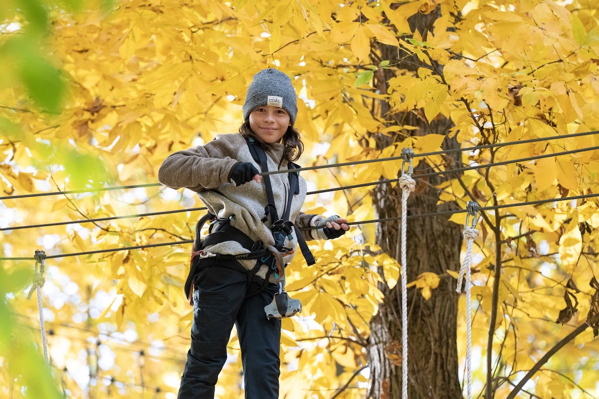 A young boy with long brown hair, wearing a gray knit "COAL" beanie and a neutral-toned fleece jacket over a black zip-up, smiles at the camera while navigating an aerial ropes course. He is wearing fingerless black gloves and is securely attached to a safety harness and belay system. He stands on a thick cable, holding onto a parallel handrail cable with both hands. The background is a vibrant, out-of-focus canopy of bright yellow autumn leaves and the thick trunk of a deciduous tree, illuminated by soft daylight.