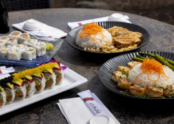 A top-down shot of a diverse spread of Japanese cuisine from Chiba in Westfield, Indiana, arranged on a dark, textured outdoor table. In the foreground, a rectangular white platter holds two specialty sushi rolls: one topped with bright yellow tobiko and avocado, and another with black tobiko and avocado. To the left, a blue glass plate features classic California rolls with crab and avocado. In the background, two dark, ribbed ceramic plates hold hot entrees: one with seared sea scallops, a mound of white rice topped with shredded carrots, and grilled asparagus; the other with a similar rice mountain and sliced teriyaki chicken. Several white napkins with black "Chiba" logos and white ceramic dipping bowls are placed around the dishes.