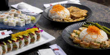 A top-down shot of a diverse spread of Japanese cuisine from Chiba in Westfield, Indiana, arranged on a dark, textured outdoor table. In the foreground, a rectangular white platter holds two specialty sushi rolls: one topped with bright yellow tobiko and avocado, and another with black tobiko and avocado. To the left, a blue glass plate features classic California rolls with crab and avocado. In the background, two dark, ribbed ceramic plates hold hot entrees: one with seared sea scallops, a mound of white rice topped with shredded carrots, and grilled asparagus; the other with a similar rice mountain and sliced teriyaki chicken. Several white napkins with black "Chiba" logos and white ceramic dipping bowls are placed around the dishes.
