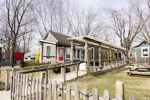 A brightly lit outdoor patio and exterior view of Chiba, a sushi restaurant in Westfield, Indiana. A light-colored wooden ramp with railings leads toward the entrance of a dark gray building with a red-framed glass door. To the right of the ramp, a long wooden pergola with horizontal beams spans the length of the walkway. The foreground features a rustic wooden picket fence decorated with a string of small lights. In the yard to the right, several wooden picnic tables are arranged on a grassy area, and bare trees are visible in the background under a pale, clear sky.