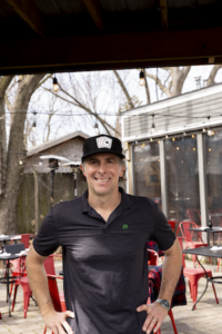 A medium shot of a smiling man, Keith Miller, the founder and owner of Chiba in Westfield, Indiana. He is wearing a black short-sleeved polo shirt and a black baseball cap with a white patch on the front. He is standing outdoors on the restaurant's patio, which features several red metal chairs and dark tables arranged on a stone-paved ground. In the background, string lights are draped between bare trees and a gray building with large windows. The overall atmosphere is bright and casual.