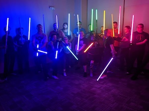 In a dimly lit room, a large group of people poses together, each holding a glowing lightsaber. The sabers illuminate the scene with a vibrant array of colors, including blue, green, yellow, pink, and red, casting colorful glows on the participants and the carpeted floor. The group is arranged in two rows, with those in the front kneeling or lunging to create a dynamic, tiered composition.
