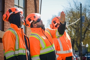 Three professional tree service workers, likely from Lumberjack Jim's Tree Service, stand together on a residential job site. They are dressed in matching high-visibility orange safety jackets and protective forestry helmets with integrated earmuffs, all looking upward as one worker points toward a tree canopy. The scene is set in front of a brick building on a bright, sunny day, emphasizing a coordinated and safety-focused team environment.