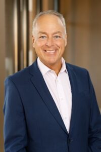 A professional headshot of a smiling middle-aged man with short, greyish-blonde hair. He is wearing a tailored navy blue blazer over a light pink or white button-down shirt. The background is a soft, out-of-focus office or corporate setting with warm, neutral tones.