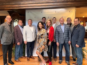 A diverse group of ten professionally dressed individuals—eight men and two women—pose for a group photo in an upscale indoor setting with dark wood paneling and a polished wood floor. They are gathered in front of a white "step and repeat" backdrop featuring a subtle, repeating logo. The group is smiling and arranged in two rows, with a woman in a floral mini dress and a woman in a red blazer at the center. The men are wearing a variety of blazers, including solid navy, grey, and patterned styles, paired with dress slacks or dark jeans.