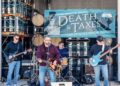 A four-piece rock band performs live on an outdoor stage at the Death & Taxes Day festival. The band consists of a lead singer and guitarist in a maroon shirt and jeans, a guitarist in a black hoodie and sunglasses, a bassist in a navy sweatshirt with a bright blue electric bass, and a drummer partially visible in the background. Behind them, several stacks of silver beer kegs and wooden barrels are arranged on metal shelving. A large teal banner hangs above the stage, featuring a top-hat-wearing skull and the text "Taxman Brewing Co. Presents DEATH & TAXES DAY - Beer Release & Tasting Festival."