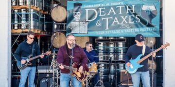 A four-piece rock band performs live on an outdoor stage at the Death & Taxes Day festival. The band consists of a lead singer and guitarist in a maroon shirt and jeans, a guitarist in a black hoodie and sunglasses, a bassist in a navy sweatshirt with a bright blue electric bass, and a drummer partially visible in the background. Behind them, several stacks of silver beer kegs and wooden barrels are arranged on metal shelving. A large teal banner hangs above the stage, featuring a top-hat-wearing skull and the text "Taxman Brewing Co. Presents DEATH & TAXES DAY - Beer Release & Tasting Festival."