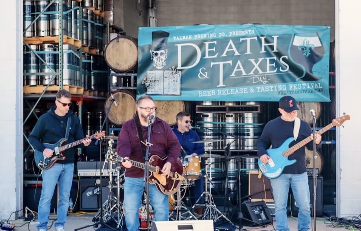 A four-piece rock band performs live on an outdoor stage at the Death & Taxes Day festival. The band consists of a lead singer and guitarist in a maroon shirt and jeans, a guitarist in a black hoodie and sunglasses, a bassist in a navy sweatshirt with a bright blue electric bass, and a drummer partially visible in the background. Behind them, several stacks of silver beer kegs and wooden barrels are arranged on metal shelving. A large teal banner hangs above the stage, featuring a top-hat-wearing skull and the text "Taxman Brewing Co. Presents DEATH & TAXES DAY - Beer Release & Tasting Festival."
