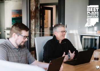 Two men sit at a light wood conference table in a modern office, engaged in a discussion with their laptops open. In the top right corner, two award logos are displayed: "Best Places to Work in Indiana 2026" and "CRN MSP 500 2026." The blackink IT logo is visible in the bottom right corner.