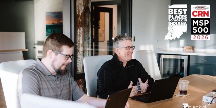 Two men sit at a light wood conference table in a modern office, engaged in a discussion with their laptops open. In the top right corner, two award logos are displayed: "Best Places to Work in Indiana 2026" and "CRN MSP 500 2026." The blackink IT logo is visible in the bottom right corner.