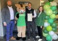 City of Noblesville Mayor Chris Jensen (center) stands with a smiling group to celebrate Ivy Tech Day 2026 at the Ivy Tech Hamilton County campus. Mayor Jensen, dressed in a black vest over a white collared shirt and green tie, holds a framed city proclamation officially declaring the day of giving. To his left is Boris the Wild Boar, the campus mascot, wearing a green "IVY TECH DAY" t-shirt. On the far left is Chuck Haberman, Assistant Director of Economic Development for the City of Noblesville and Chair of the Ivy Tech Campus Board of Trustees, wearing a gray blazer and blue trousers. On the far right is Rachel Kartz, Chancellor of the Ivy Tech Hamilton County campus, dressed in a green blazer and black pants. A tall decorative column of green and white balloons stands next to Chancellor Kartz against the modern office backdrop.