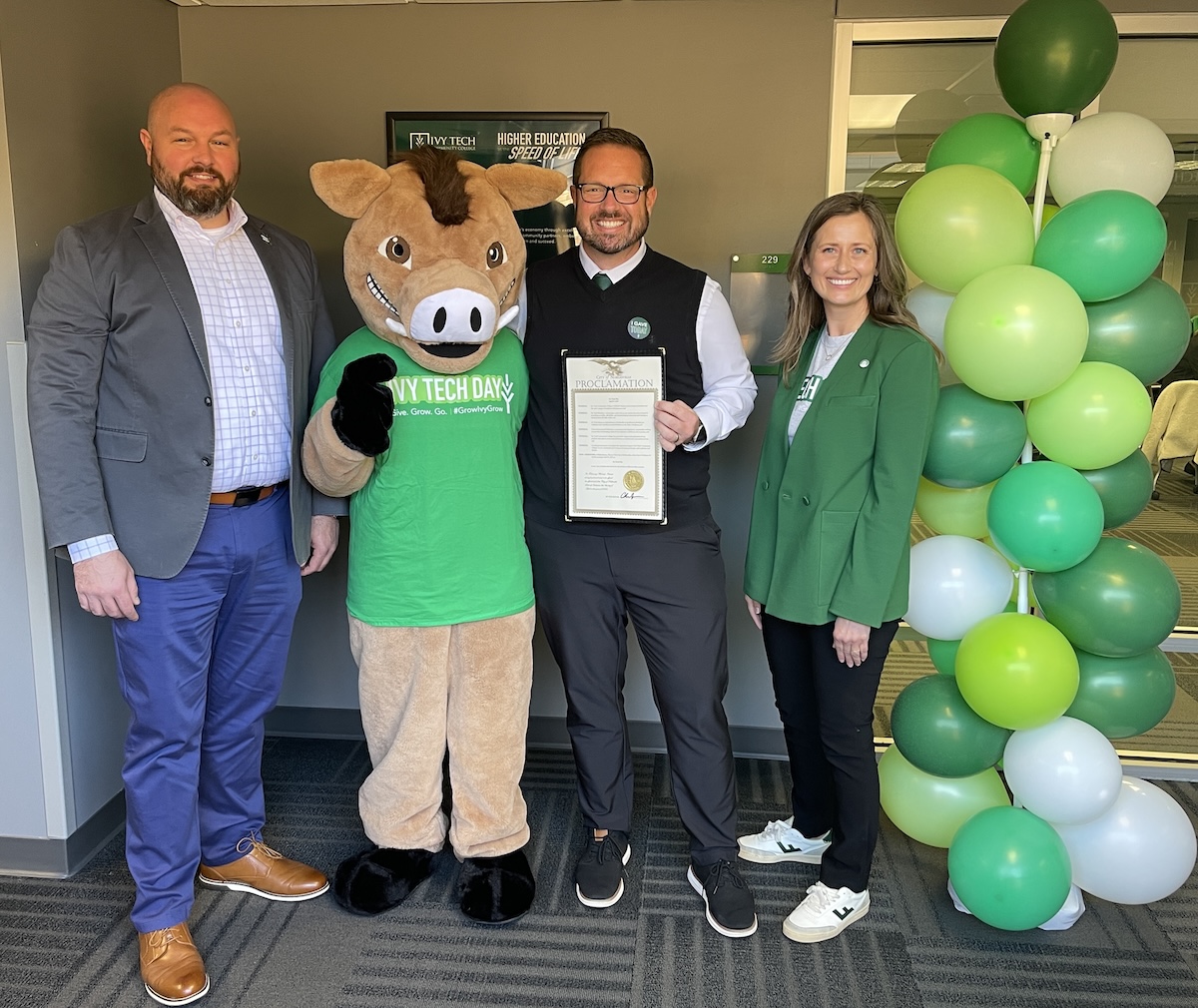 City of Noblesville Mayor Chris Jensen (center) stands with a smiling group to celebrate Ivy Tech Day 2026 at the Ivy Tech Hamilton County campus. Mayor Jensen, dressed in a black vest over a white collared shirt and green tie, holds a framed city proclamation officially declaring the day of giving. To his left is Boris the Wild Boar, the campus mascot, wearing a green "IVY TECH DAY" t-shirt. On the far left is Chuck Haberman, Assistant Director of Economic Development for the City of Noblesville and Chair of the Ivy Tech Campus Board of Trustees, wearing a gray blazer and blue trousers. On the far right is Rachel Kartz, Chancellor of the Ivy Tech Hamilton County campus, dressed in a green blazer and black pants. A tall decorative column of green and white balloons stands next to Chancellor Kartz against the modern office backdrop.