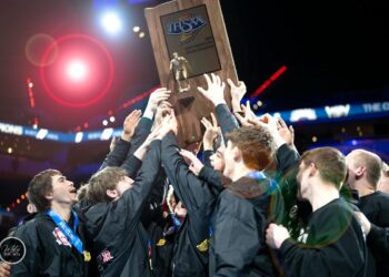 A triumphant group of teenage boys, members of the Center Grove Trojans wrestling team, celebrate their victory at the 2026 IHSAA Boys Wrestling State Finals. They are huddled together in their black team jackets, several wearing state finalist medals around their necks, as they collectively hoist a large wooden championship trophy into the air. The trophy features a golden wrestler figurine and the IHSAA logo. The scene is set inside the brightly lit Gainbridge Fieldhouse arena in Indianapolis, with a soft-focus background of the cheering crowd and stadium lights.
