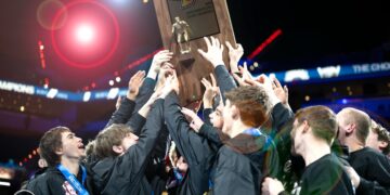 A triumphant group of teenage boys, members of the Center Grove Trojans wrestling team, celebrate their victory at the 2026 IHSAA Boys Wrestling State Finals. They are huddled together in their black team jackets, several wearing state finalist medals around their necks, as they collectively hoist a large wooden championship trophy into the air. The trophy features a golden wrestler figurine and the IHSAA logo. The scene is set inside the brightly lit Gainbridge Fieldhouse arena in Indianapolis, with a soft-focus background of the cheering crowd and stadium lights.