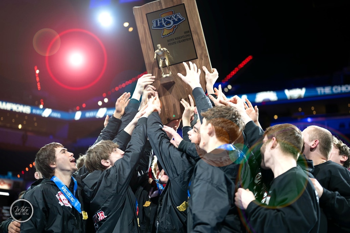 A triumphant group of teenage boys, members of the Center Grove Trojans wrestling team, celebrate their victory at the 2026 IHSAA Boys Wrestling State Finals. They are huddled together in their black team jackets, several wearing state finalist medals around their necks, as they collectively hoist a large wooden championship trophy into the air. The trophy features a golden wrestler figurine and the IHSAA logo. The scene is set inside the brightly lit Gainbridge Fieldhouse arena in Indianapolis, with a soft-focus background of the cheering crowd and stadium lights.