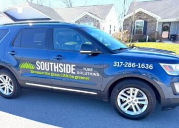 A side profile of a dark blue Ford Explorer parked on a residential street in front of modern suburban homes. The front door of the SUV features a professional vinyl decal with the "Southside Turf Solutions" logo, which includes a stylized green grass graphic and the slogan "Because the grass CAN be greener." The company's phone number, 317-286-1636, is clearly displayed in white text on the front fender. The vehicle has chrome accents along the bottom and multi-spoke silver alloy wheels.