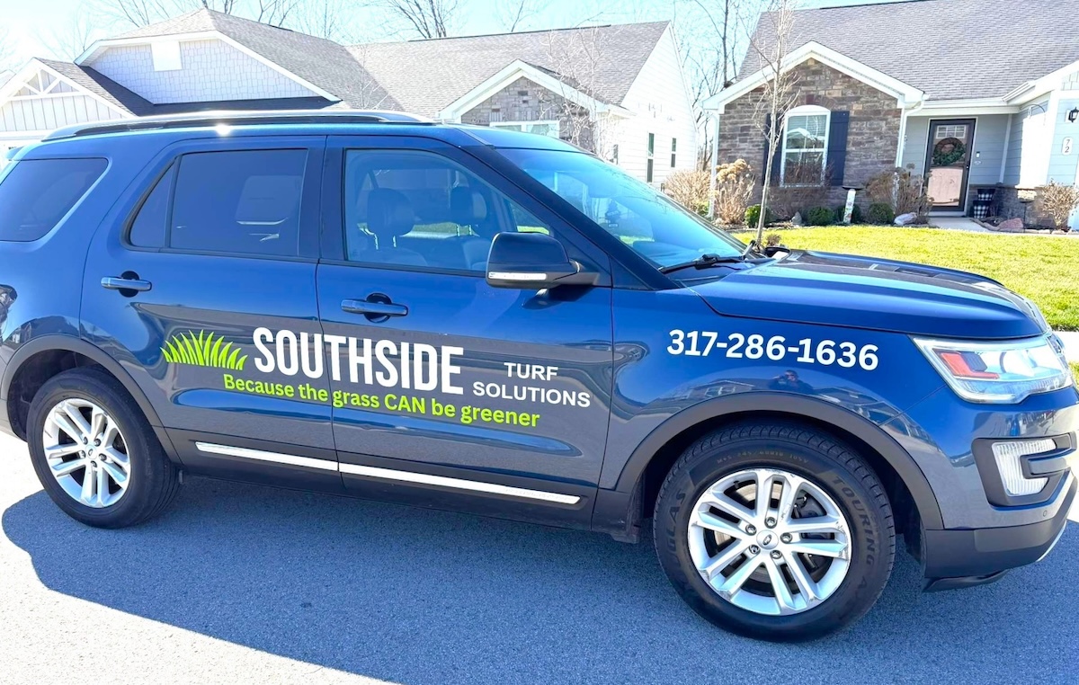 A side profile of a dark blue Ford Explorer parked on a residential street in front of modern suburban homes. The front door of the SUV features a professional vinyl decal with the "Southside Turf Solutions" logo, which includes a stylized green grass graphic and the slogan "Because the grass CAN be greener." The company's phone number, 317-286-1636, is clearly displayed in white text on the front fender. The vehicle has chrome accents along the bottom and multi-spoke silver alloy wheels.