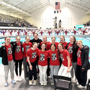 Twelve members of the Center Grove High School girls' swimming and diving team stand together for a group photo at the IU Natatorium in Indianapolis. They are positioned in front of an Olympic-sized swimming pool, with rows of empty spectator bleachers rising behind them. Most of the athletes are wearing matching red t-shirts that feature a white outline of Indiana and text celebrating their status as 2026 IHSAA State Finalists. They are smiling and have their arms around each other's shoulders. Above the pool, a large American flag and various regional banners are suspended from the high, industrial-style ceiling.