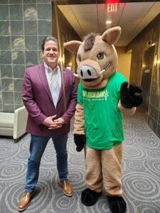A full-length photo shows a man in a maroon blazer and jeans standing next to the Ivy Tech Hamilton County mascot, Boris the Wild Boar, in a modern lobby. The man is smiling at the camera with his hands clasped in front of him, wearing light brown leather shoes. Boris, the large tan boar mascot with black hooves and small white tusks, is wearing a bright green t-shirt with the "IVY TECH DAY" logo and a white tree graphic. He is gesturing with one black-gloved hand. The room features a gray patterned carpet, a tiled accent wall on the left, and a hallway with an illuminated "EXIT" sign in the background.