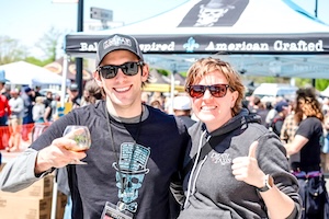 A medium shot shows a man and a woman smiling for a photo at an outdoor festival. The man, on the left, is wearing a black t-shirt with a skeleton in a top hat—the Taxman Brewing logo—and a matching black baseball cap. He holds up a clear snifter glass of dark beer. The woman, to his right, is wearing a dark gray zip-up hoodie and dark sunglasses, giving a thumbs-up to the camera. They are standing in front of a white event tent with a banner that reads "Belgian Inspired - American Crafted" and features the same skull logo. A large, blurred crowd and other festival tents are visible in the background under a bright, sunny sky