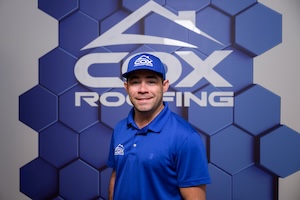 A professional headshot of a smiling man wearing a royal blue polo shirt and a matching baseball cap, both featuring the Cox Roofing logo. He is standing in front of a blue and grey accent wall with a large, modern 3D honeycomb pattern and the prominent white Cox Roofing logo at the top.