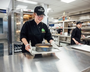 A smiling young woman in a black chef's coat and a black baseball cap with the "FOODIES ROCK" logo carefully pulls a fresh cake in a springform pan out of an industrial oven. She is using a white kitchen towel to protect her hands as she places the cake onto a metal baking sheet on a stainless steel counter. In the background, another student in a black chef's coat works at a different station. The commercial kitchen is brightly lit and features open metal shelving stocked with white dishes and supplies.