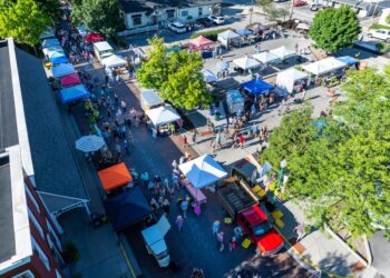 An aerial, high-angle view captures a bustling outdoor market in Zionsville, Indiana. Dozens of white and colorful vendor tents are arranged in rows along a brick-paved street and an adjacent parking area. A diverse crowd of people is seen browsing the various stalls under a bright, sunny sky. A red pickup truck is parked in the foreground near the market entrance. Mature green trees are scattered throughout the space, providing shade for shoppers. In the bottom left corner, a portion of a red brick building with a sign ending in "SQUARE" is visible.