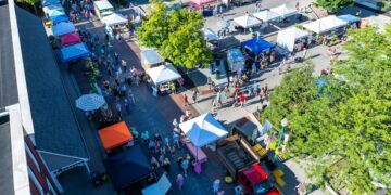 An aerial, high-angle view captures a bustling outdoor market in Zionsville, Indiana. Dozens of white and colorful vendor tents are arranged in rows along a brick-paved street and an adjacent parking area. A diverse crowd of people is seen browsing the various stalls under a bright, sunny sky. A red pickup truck is parked in the foreground near the market entrance. Mature green trees are scattered throughout the space, providing shade for shoppers. In the bottom left corner, a portion of a red brick building with a sign ending in "SQUARE" is visible.