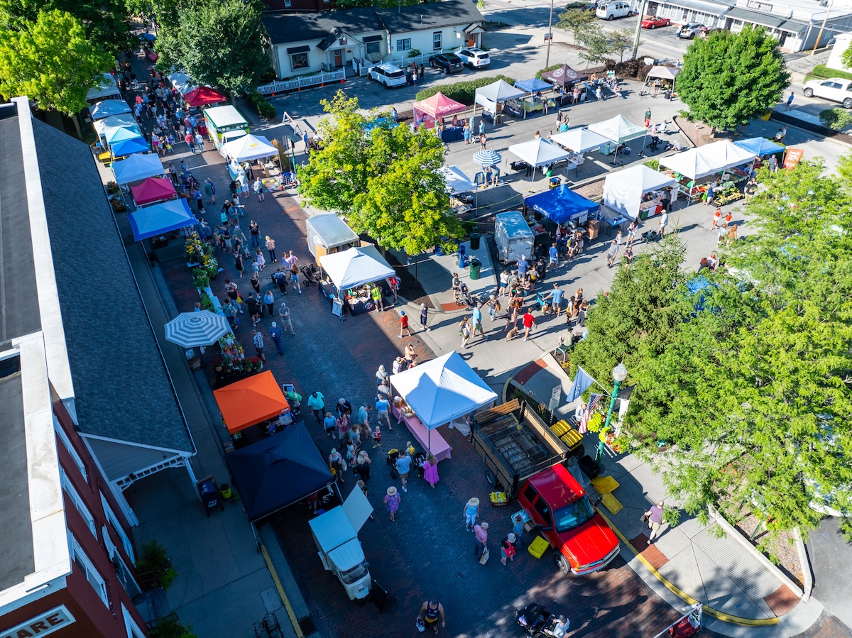 An aerial, high-angle view captures a bustling outdoor market in Zionsville, Indiana. Dozens of white and colorful vendor tents are arranged in rows along a brick-paved street and an adjacent parking area. A diverse crowd of people is seen browsing the various stalls under a bright, sunny sky. A red pickup truck is parked in the foreground near the market entrance. Mature green trees are scattered throughout the space, providing shade for shoppers. In the bottom left corner, a portion of a red brick building with a sign ending in "SQUARE" is visible.
