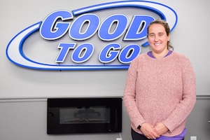 A professional headshot of Nikki Earp, a member of the executive leadership team at Good Oil Company. She is smiling warmly, wearing a textured, pink heather-knit sweater and a black smartwatch, standing in front of a blue and silver "GOOD TO GO" company logo. A modern black electric fireplace is mounted on the grey wall behind her.