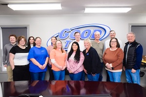 A group of 13 employees from Good Oil Company stand together and smile for a team photo in a modern office conference room. They are positioned in front of a large, blue and silver "GOOD TO GO" company logo mounted on the back wall. The staff is dressed in a mix of business casual and professional attire, including blue and orange company-branded tops, set against a clean white wall with bright overhead lighting.
