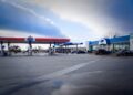 A wide-angle view of a modern Amoco gas station and travel center under a cloudy sky. The facility features two large red, white, and blue fueling canopies and a primary convenience store building. The storefront prominently displays logos for Coen Markets, Coen Kitchen, and Subway, indicating a variety of dining and shopping options on-site. Several vehicles are parked in the spacious, paved lot in front of the blue-roofed building.