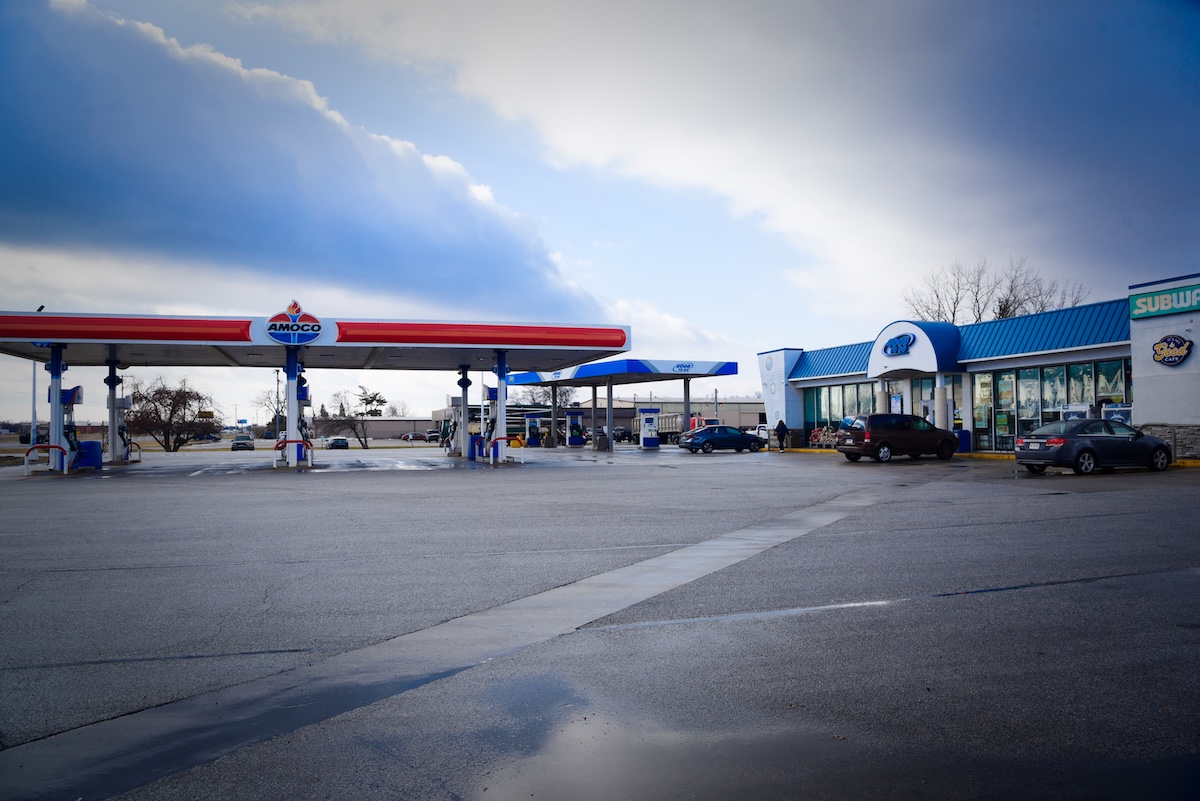 A wide-angle view of a modern Amoco gas station and travel center under a cloudy sky. The facility features two large red, white, and blue fueling canopies and a primary convenience store building. The storefront prominently displays logos for Coen Markets, Coen Kitchen, and Subway, indicating a variety of dining and shopping options on-site. Several vehicles are parked in the spacious, paved lot in front of the blue-roofed building.