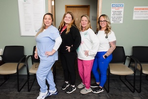 Four women from a professional medical or administrative team stand together in a healthcare facility's waiting area. They are dressed in various professional attire, including light blue scrubs, a black zip-up jacket, a white long-sleeved shirt with pink scrubs, and a white t-shirt with cobalt blue scrubs. The scene is set in a neutral-toned room with comfortable waiting room chairs and official notice posters on the wall.