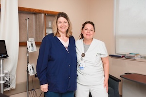 wo healthcare professionals, a nurse and a staff member, stand together and smile in a clinical hospital room. The woman on the left is wearing a navy blue button-front cardigan over a light-colored top, while the nurse on the right is in white medical scrubs with a visible ID badge. In the background, a medical pole with an infusion pump is positioned near a white hospital curtain.
Navy Blue Cardigan
