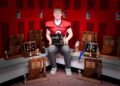 A professional portrait of Myles McLaughlin, a standout football player for the Knox High School Redskins. He is sitting in a locker room, wearing his red and black #5 jersey, surrounded by a large collection of IHSAA sectional and regional championship trophies. He holds a "Hoosier North Athletic Conference" award, representing his record-breaking career as Indiana’s all-time leading rusher.