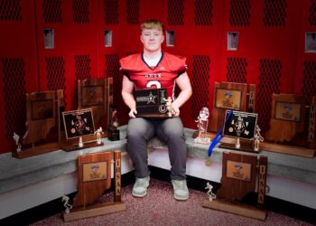 A professional portrait of Myles McLaughlin, a standout football player for the Knox High School Redskins. He is sitting in a locker room, wearing his red and black #5 jersey, surrounded by a large collection of IHSAA sectional and regional championship trophies. He holds a "Hoosier North Athletic Conference" award, representing his record-breaking career as Indiana’s all-time leading rusher.