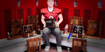 A professional portrait of Myles McLaughlin, a standout football player for the Knox High School Redskins. He is sitting in a locker room, wearing his red and black #5 jersey, surrounded by a large collection of IHSAA sectional and regional championship trophies. He holds a "Hoosier North Athletic Conference" award, representing his record-breaking career as Indiana’s all-time leading rusher.