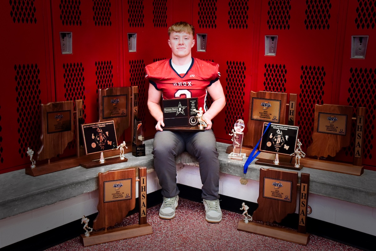 A professional portrait of Myles McLaughlin, a standout football player for the Knox High School Redskins. He is sitting in a locker room, wearing his red and black #5 jersey, surrounded by a large collection of IHSAA sectional and regional championship trophies. He holds a "Hoosier North Athletic Conference" award, representing his record-breaking career as Indiana’s all-time leading rusher.