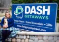 A woman sits on a stone wall next to a large blue sign for "DASH GETAWAYS." The sign lists "Group Tours • Travel Essentials • Gifts" and the website dashgetawaytravel.com. She is wearing a navy blue sweater and is wrapped in a green and blue plaid blanket while holding a plush otter toy.