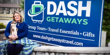 A woman sits on a stone wall next to a large blue sign for "DASH GETAWAYS." The sign lists "Group Tours • Travel Essentials • Gifts" and the website dashgetawaytravel.com. She is wearing a navy blue sweater and is wrapped in a green and blue plaid blanket while holding a plush otter toy.