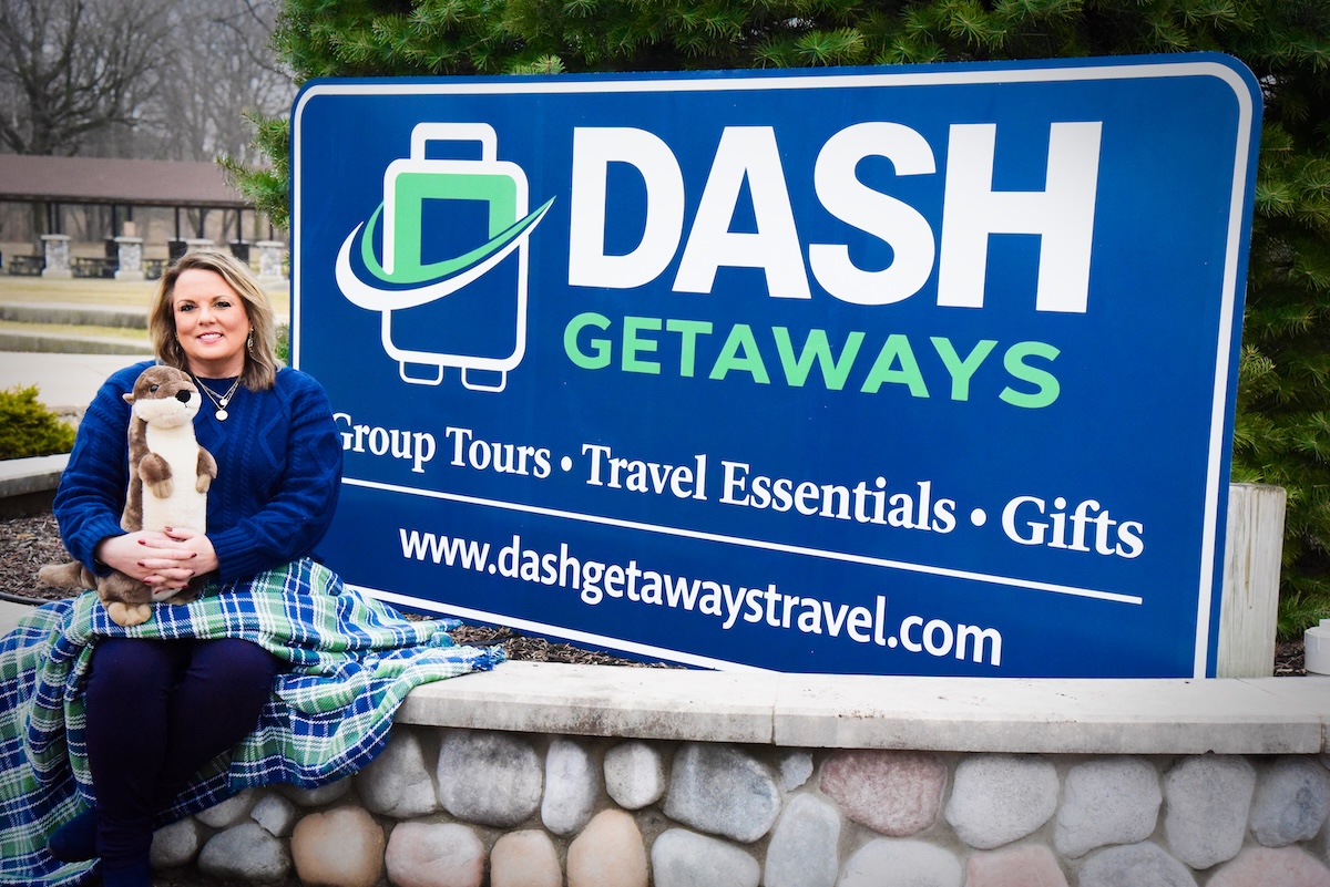 A woman sits on a stone wall next to a large blue sign for "DASH GETAWAYS." The sign lists "Group Tours • Travel Essentials • Gifts" and the website dashgetawaytravel.com. She is wearing a navy blue sweater and is wrapped in a green and blue plaid blanket while holding a plush otter toy.