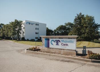 The entrance to Majestic Care of Carmel features a large, rectangular monument sign in the foreground with a blue heart-shaped logo and the facility's name in bold blue and red lettering. In the background, a multi-story white building with the same logo stands prominently behind a manicured green lawn and mature trees. A paved driveway leads toward the building under a clear, bright blue sky.