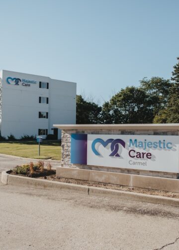 The entrance to Majestic Care of Carmel features a large, rectangular monument sign in the foreground with a blue heart-shaped logo and the facility's name in bold blue and red lettering. In the background, a multi-story white building with the same logo stands prominently behind a manicured green lawn and mature trees. A paved driveway leads toward the building under a clear, bright blue sky.