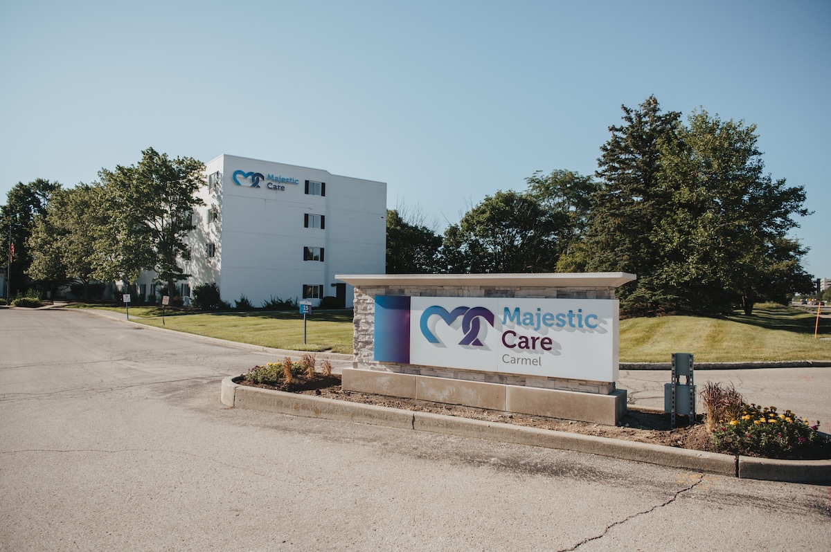 The entrance to Majestic Care of Carmel features a large, rectangular monument sign in the foreground with a blue heart-shaped logo and the facility's name in bold blue and red lettering. In the background, a multi-story white building with the same logo stands prominently behind a manicured green lawn and mature trees. A paved driveway leads toward the building under a clear, bright blue sky.