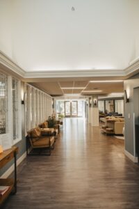 A long, brightly lit hallway in a modern senior living facility features dark wood-look flooring and neutral grey and white walls. To the left, a row of decorative stained-glass windows lines the corridor, with several tan leather armchairs and small wooden side tables providing a seating area. To the right, an open living space with plush sofas and a console table is visible. Sleek, black wall sconces and recessed ceiling lights illuminate the space, which leads toward a pair of glass double doors at the far end of the hallway.