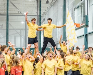 A high-energy celebration takes place at Westfield High School for the 2026 Dance Marathon. Dozens of students, all wearing matching bright yellow "WHSDM" t-shirts, are cheering and jumping with their arms raised. In the center, two young men are being hoisted above the crowd; one is punching the air in triumph, while the other holds a large yellow and white flag. The event is held in an indoor athletic facility with glass partitions and a basketball hoop visible on the right. The room is filled with a sense of pure joy and accomplishment. 