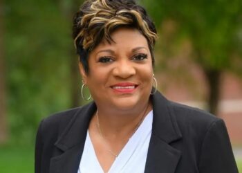 A professional studio portrait of Deb Whitfield, the Mayor of Lawrence, Indiana. She is a Black woman with a warm, confident smile and short, styled dark hair with golden-blonde highlights. She is wearing a black blazer over a white draped-neck blouse, accented by silver hoop earrings and a delicate silver necklace. The background is a soft-focus green landscape with trees, suggesting an outdoor setting with even, natural lighting.